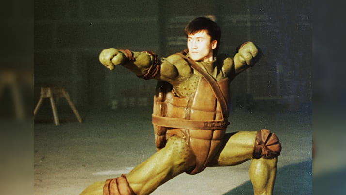 Hoyoung Pak crouches in a martial arts pose with his arm extended, on the set of the Teenage Mutant Ninja Turtles film He wears a turtle-suit costume, complete with a shell on his back.