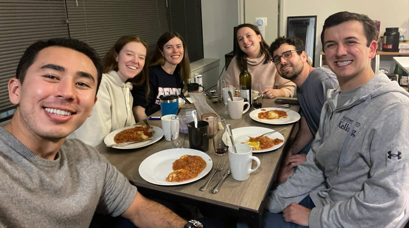 Group of six Kellogg students smiling at the camera while seated around a dinner table, enjoying plates of pasta and drinks. One person wears a Northwestern Kellogg sweatshirt. The setting appears casual and friendly, emphasizing community and camaraderie.