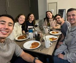 Group of six Kellogg students smiling at the camera while seated around a dinner table, enjoying plates of pasta and drinks. One person wears a Northwestern Kellogg sweatshirt. The setting appears casual and friendly, emphasizing community and camaraderie.