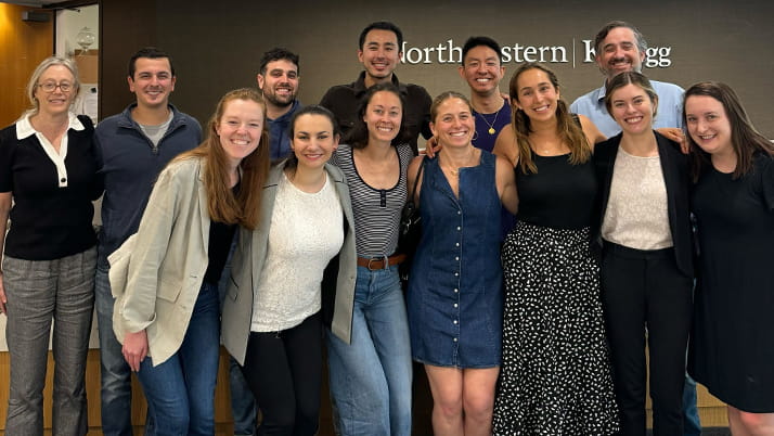 Group of thirteen Kellogg students and faculty standing and smiling together in front of a wall with the Northwestern Kellogg logo. The group appears cheerful and united, reflecting a strong sense of community.
