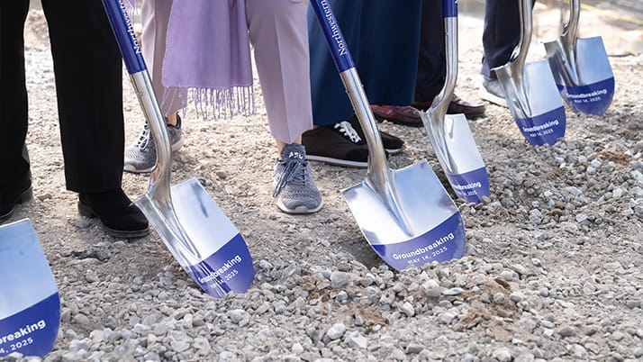 Participants in the groundbreaking ceremony stand in a row and hold silver and purple ceremonial shovels.