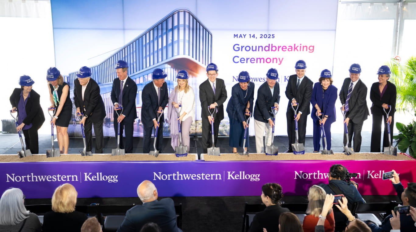A group of people wearing purple Kellogg hard hats pose with shovels during a groundbreaking ceremony for a new Northwestern Kellogg building on May 14, 2025. A large backdrop shows a rendering of the future structure, and a purple banner with the Northwestern Kellogg logo spans the front of the stage.