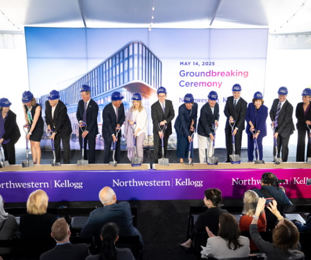 A group of people wearing purple Kellogg hard hats pose with shovels during a groundbreaking ceremony for a new Northwestern Kellogg building on May 14, 2025. A large backdrop shows a rendering of the future structure, and a purple banner with the Northwestern Kellogg logo spans the front of the stage.