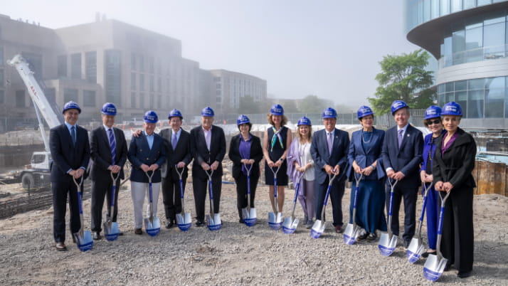 Thirteen people wearing purple Kellogg hard hats and holding shovels stand on a gravel construction site during the May 14, 2025, groundbreaking ceremony for a new Northwestern Kellogg building. The current Global Hub is visible at right.