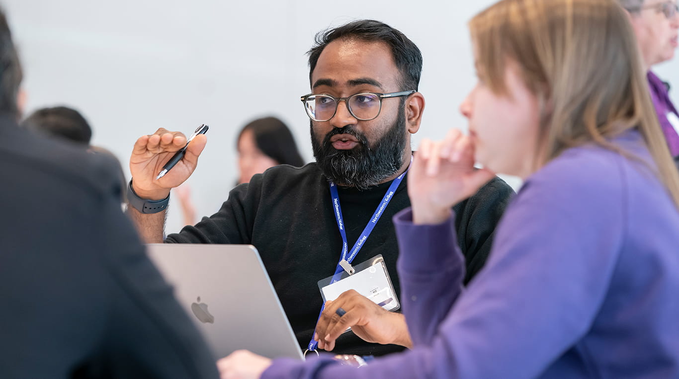 A Kellogg student wearing a Northwestern lanyard gestures while speaking during a group discussion. A laptop is open in front of him, and other students are seated nearby, engaged in conversation.