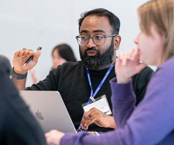 A Kellogg student wearing a Northwestern lanyard gestures while speaking during a group discussion. A laptop is open in front of him, and other students are seated nearby, engaged in conversation.