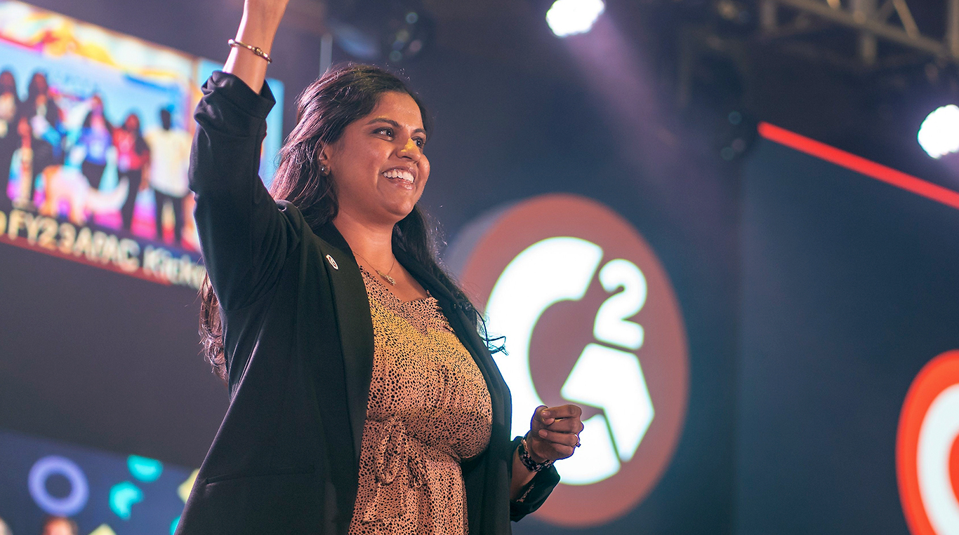 Kellogg alumna Priti Patel stands on stage smiling and raising her hand in celebration during a corporate event. The G2 company logo is visible on a large screen in the background.