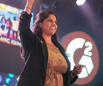 Kellogg alumna Priti Patel stands on stage smiling and raising her hand in celebration during a corporate event. The G2 company logo is visible on a large screen in the background.