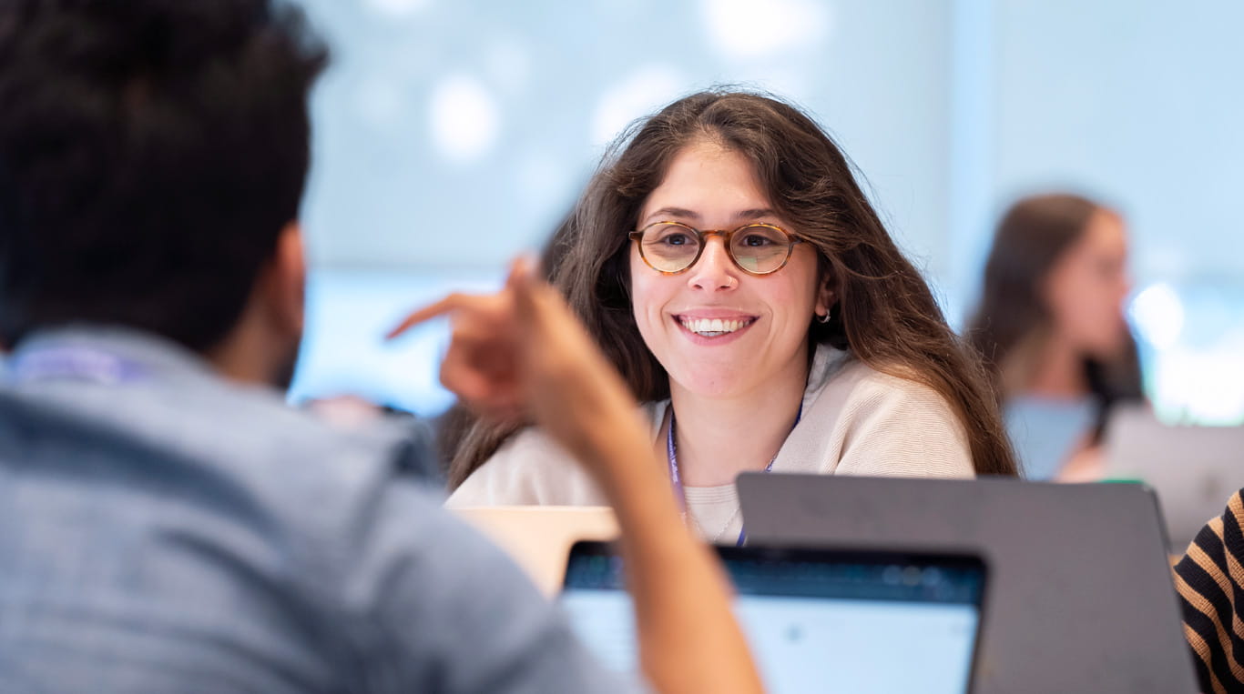 A smiling Kellogg student listens attentively to a classmate during a collaborative classroom discussion. Open laptops are visible on the table, suggesting an interactive, tech-enabled learning environment.