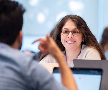 A smiling Kellogg student listens attentively to a classmate during a collaborative classroom discussion. Open laptops are visible on the table, suggesting an interactive, tech-enabled learning environment.