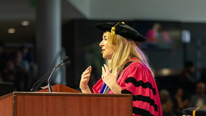 Dean Francesca Cornelli wears a red and black robe and black cap as she delivers her address at Convocation 2025.