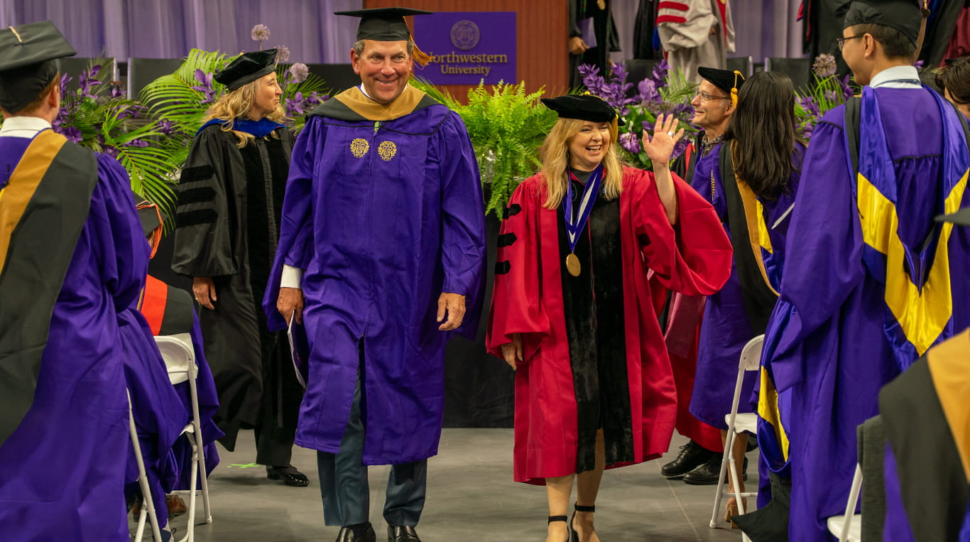Speaker John Schlifske and Dean Francesca Cornelli wear graduation regalia and march out of the 2025 Kellogg convocation ceremony, smiling and waving at graduates.
