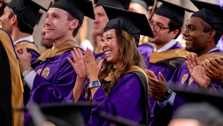 Kellogg graduates wearing purple gowns and black caps smile and applaud during the EMBA convocation ceremony.