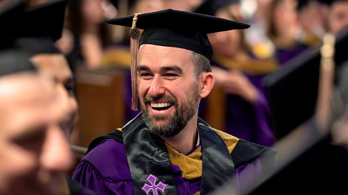 A Kellogg graduate wearing a purple gown and black cap turns and smiles to a classmate during the EMBA convocation ceremony.