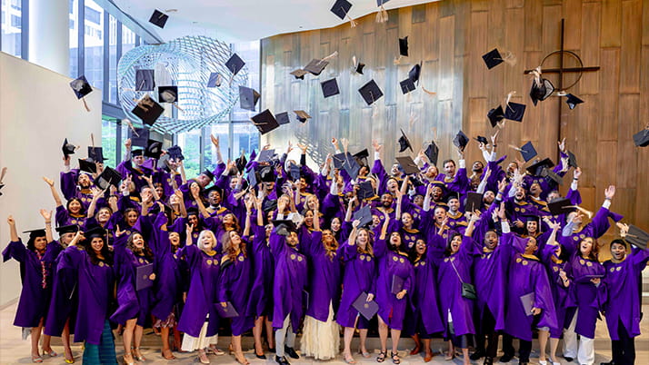 The EMBA Class of 2024 poses for a group photo, all wearing purple robes and throwing their graduation caps into the air.
