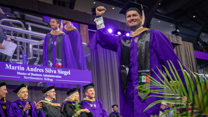A Kellogg graduate wearing a purple robe and black cap celebrates by raising his hand in the air while walking across the stage at convocation.