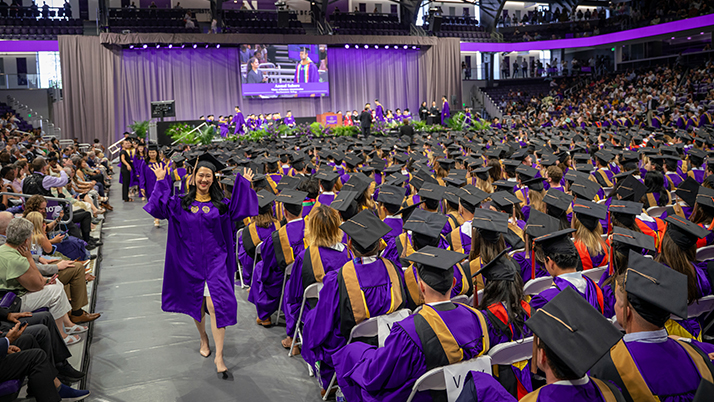 A wide photo of Ryan Arena shows the audience of Kellogg Class of 2025 graduates. A new graduate celebrates while walking down a side aisle.