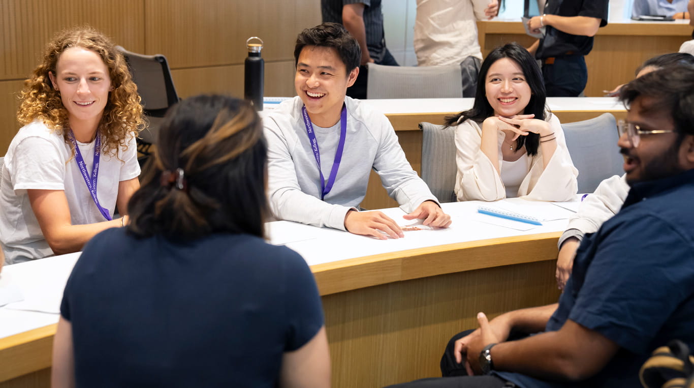 Two Kellogg students smile and engage in conversation during a classroom session. They are seated at a curved table with notebooks and nameplates in front of them, suggesting a collaborative learning environment.