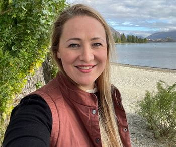 A woman in a black long sleeve shirt and red vest with a lake behind her
