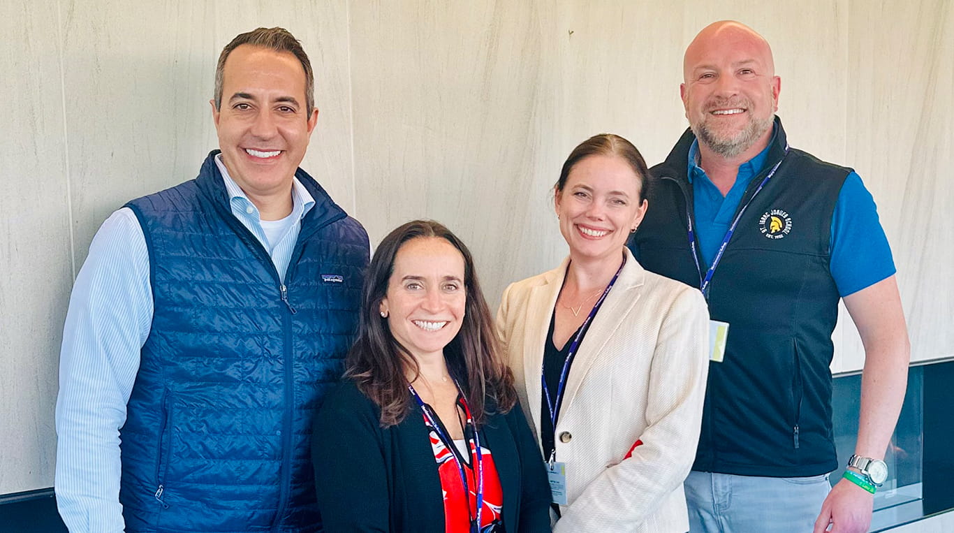 Four Executive Education participants stand together and smile at the camera during a Kellogg program session, wearing name badges and lanyards.