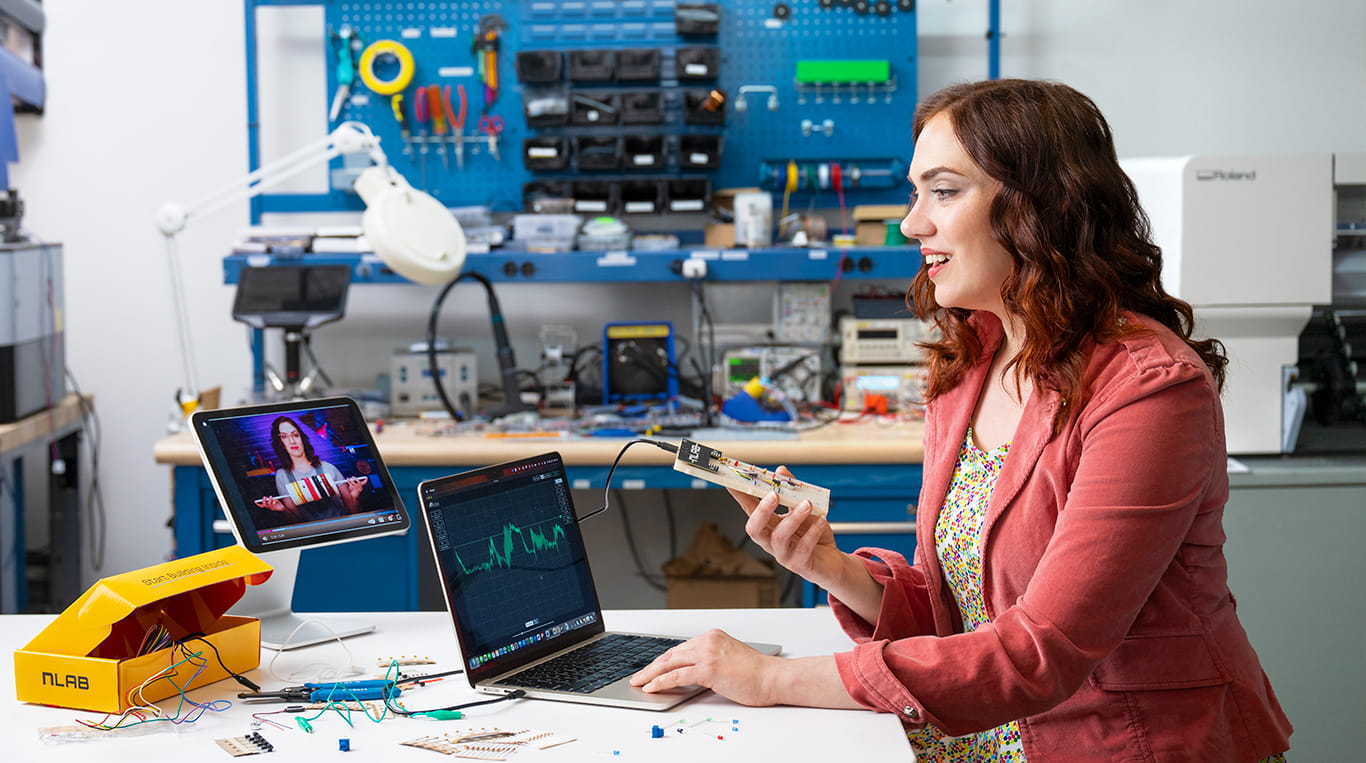 Kellogg student Angie Mercurio sits at a workbench in a lab, smiling as she tests a circuit board connected to her laptop. A video tutorial featuring the same woman plays on a nearby screen, and electronic tools and components are spread across the desk.