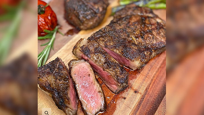 A juicy, freshly grilled steak is displayed on a cutting board, surrounded by fresh herbs and cooked vegetables.