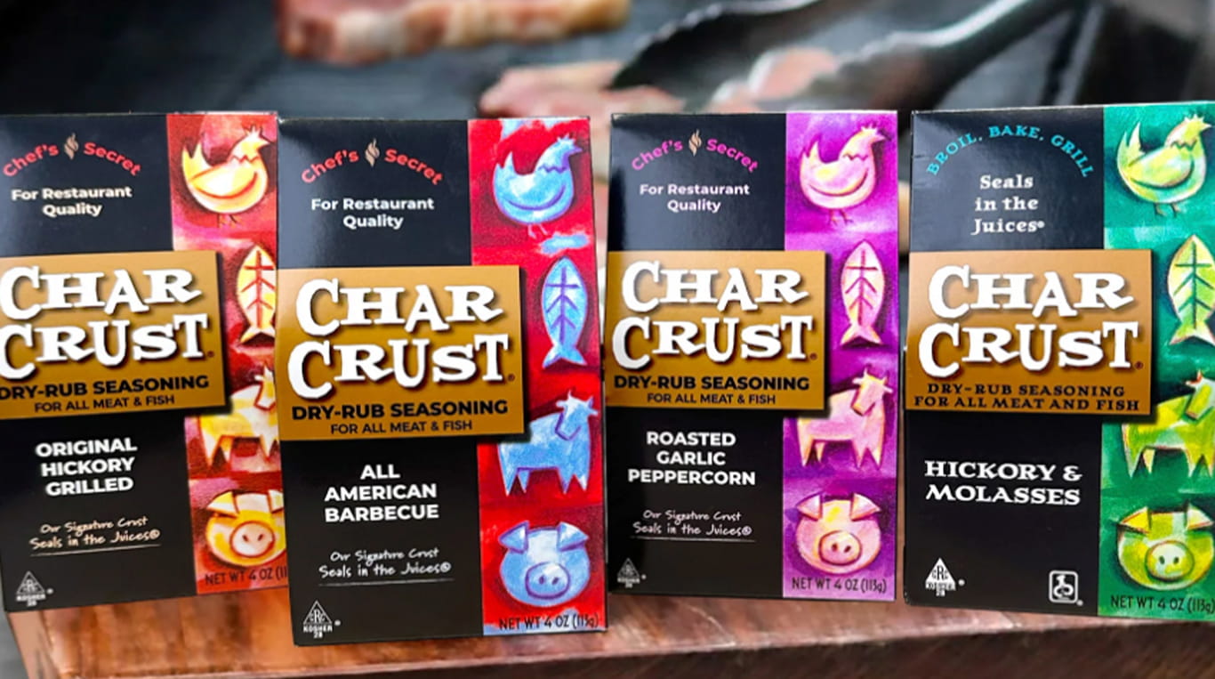A row of Char Crust dry-rub seasoning boxes is displayed on a table in front of a grill. Each box features bold, colorful artwork and flavors including Original Hickory Grilled, All American Barbecue, Roasted Garlic Peppercorn, and Hickory & Molasses.