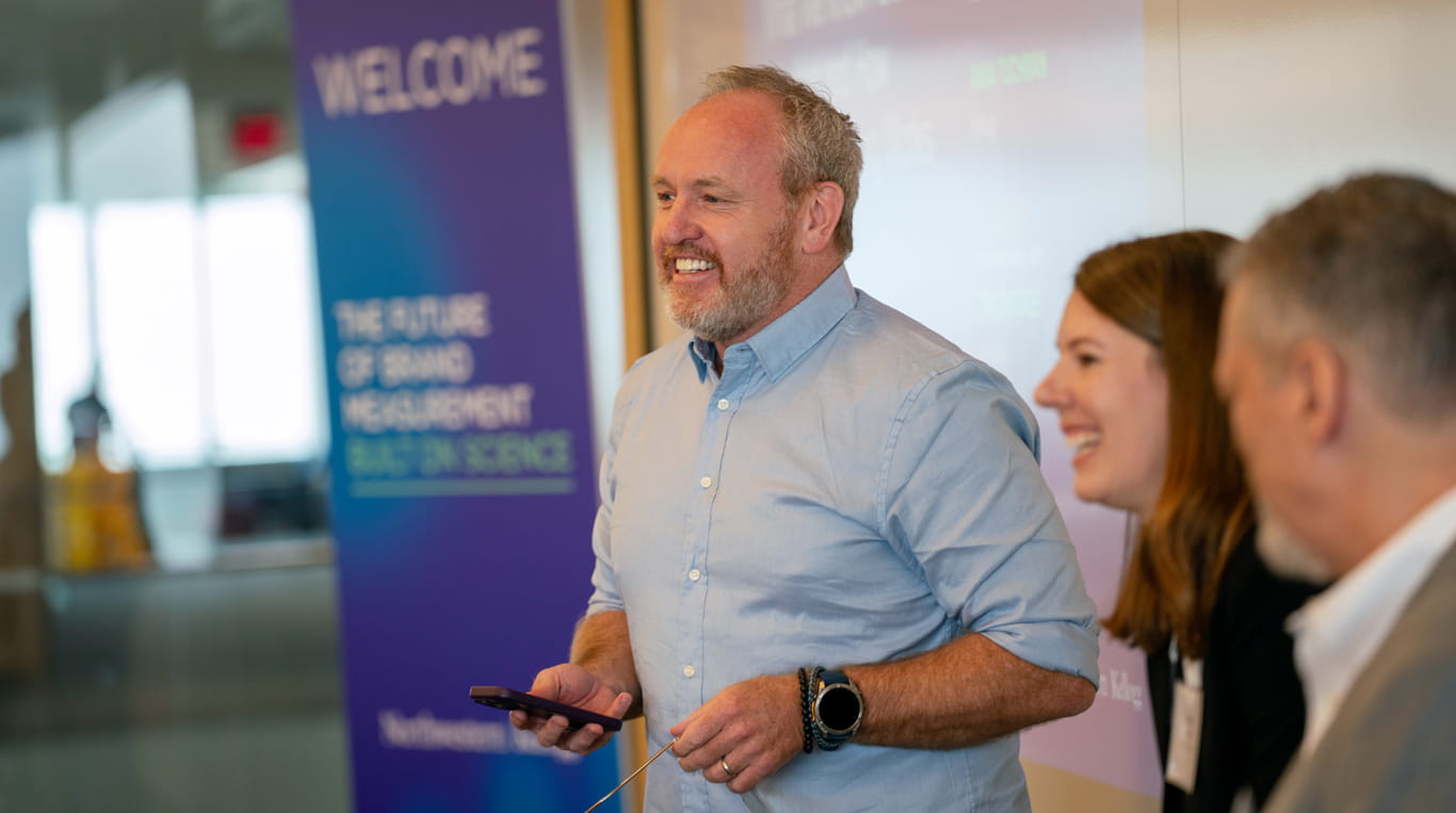 A smiling speaker holds a phone while presenting at the Northwestern Kellogg Brand Measurement Conference, with a welcome sign and other panelists visible in the background.