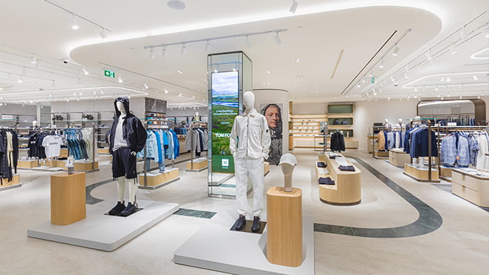 Interior of a Harry Rosen store. A modern, high-end men’s clothing store with sleek lighting and neutral tones features mannequins dressed in casual wear, digital signage, and neatly arranged racks of stylish apparel.