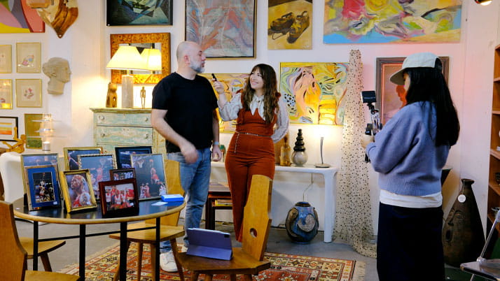 A woman interviewing a man while another woman records them in a colorfully decorated room.