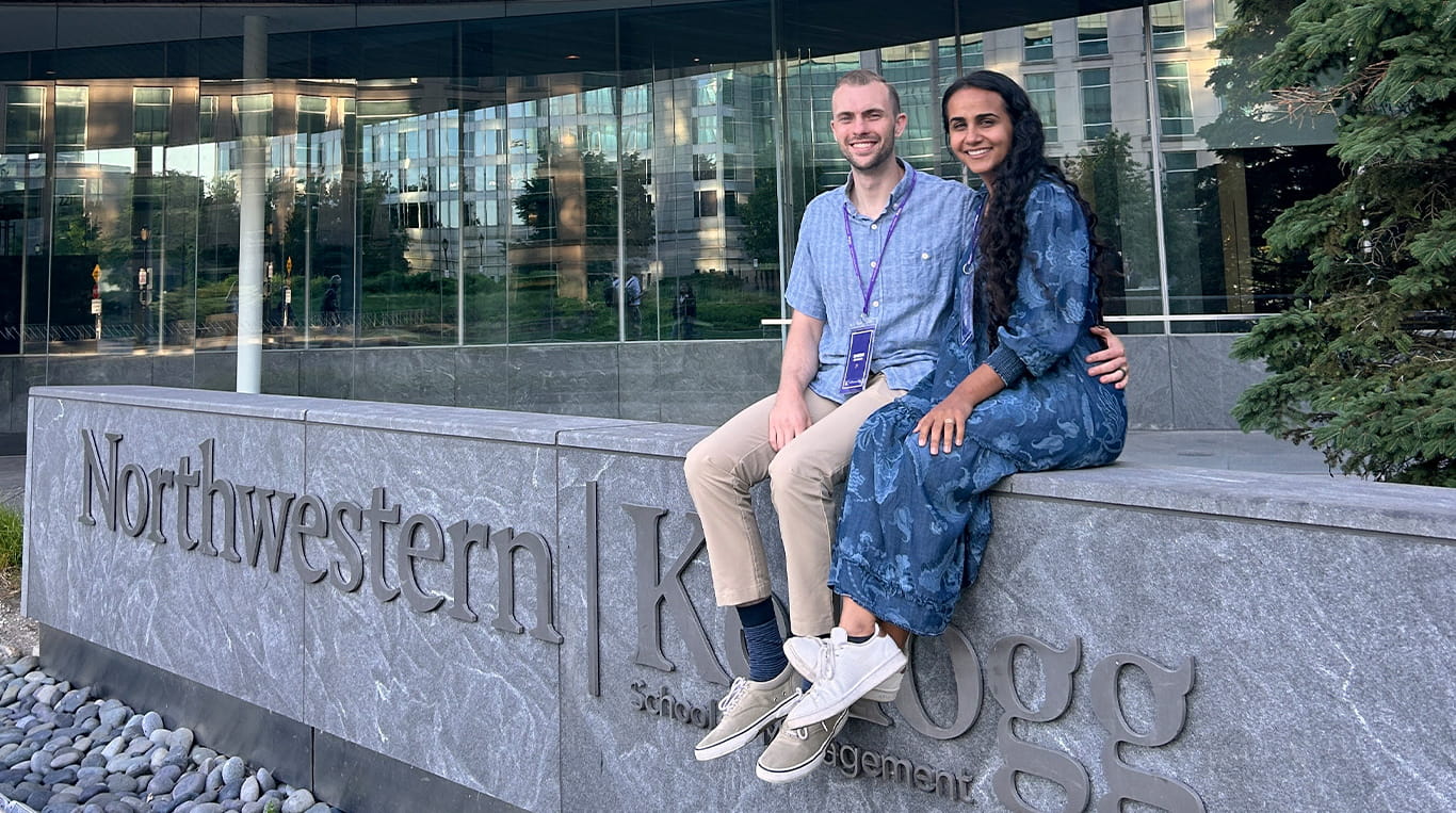 Two people sit together on the stone sign for Northwestern Kellogg School of Management, smiling at the camera. The glass-fronted Global Hub building and greenery are visible in the background.