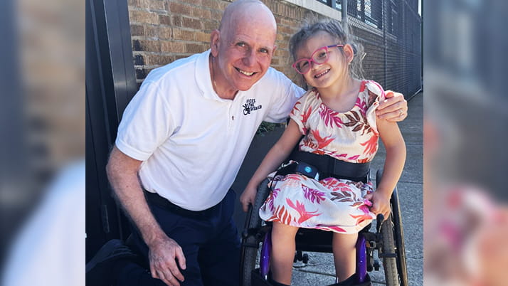 An older man in a white polo shirt kneels beside a young girl with glasses who is seated in a purple wheelchair, both smiling warmly at the camera. 