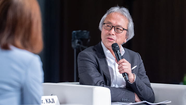 An Asian man with white hair wearing a sport coat and holding a mic