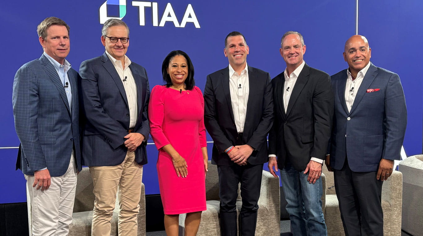 A group of six business professionals pose together in front of a blue backdrop with the TIAA logo. Kourtney Gibson ’10 MBA stands in the center wearing a bright pink dress, while the men are dressed in suits or blazers, all smiling toward the camera.