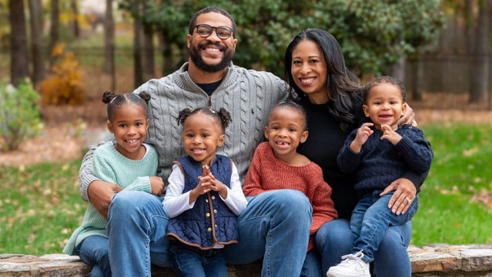 A smiling family of six poses outdoors on a stone bench. The parents sit in the back with their arms around their four young children, all dressed in sweaters and jeans, with autumn trees and greenery in the background.