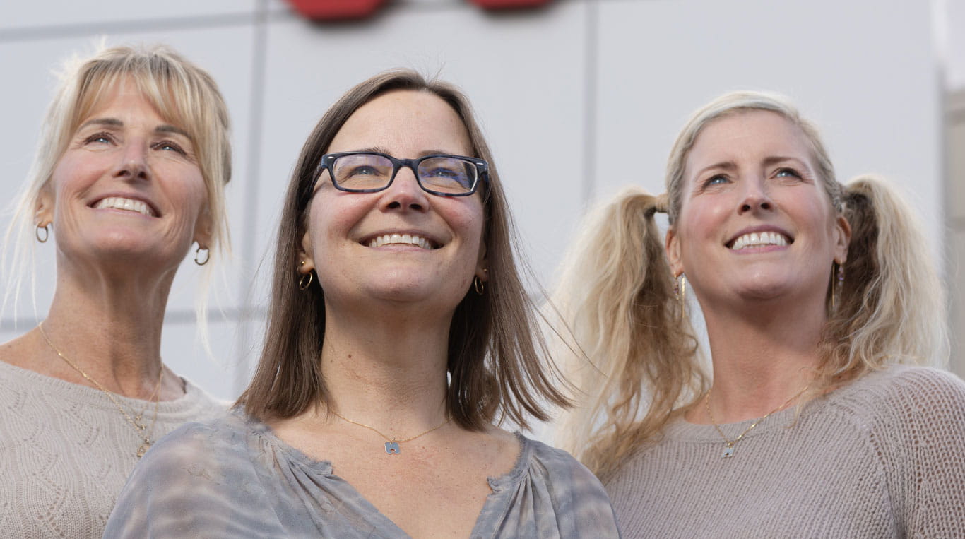 Sisters Stephanie Jackson, Kimberly Paxton-Hanger and Melissa Steiner stand together outside in front of Kwik Lok headquarters. They are looking upward, wearing casual tops and light jewelry, with sunlight highlighting their faces.