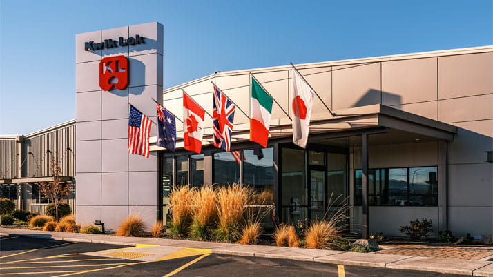 The exterior of a modern industrial building with a "Kwik Lok" sign and logo on a tall pillar. Several international flags, including those of the United States, Australia, Canada, the United Kingdom, Italy, and Japan, are displayed above the entrance.