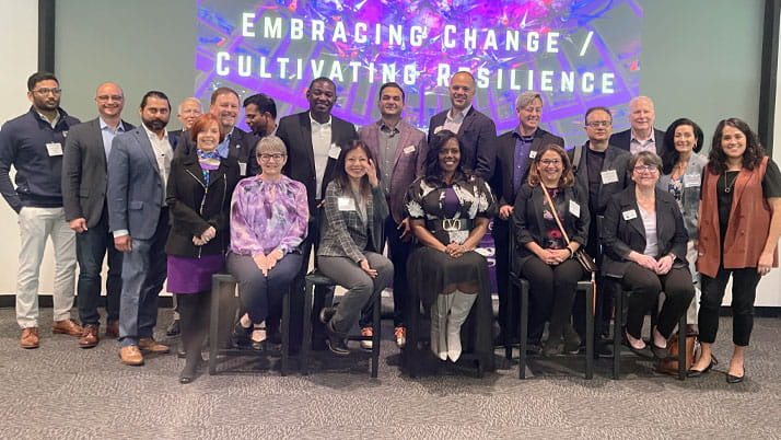 Five women sitting in chairs surrounded by men and women during a professional panel discussion.