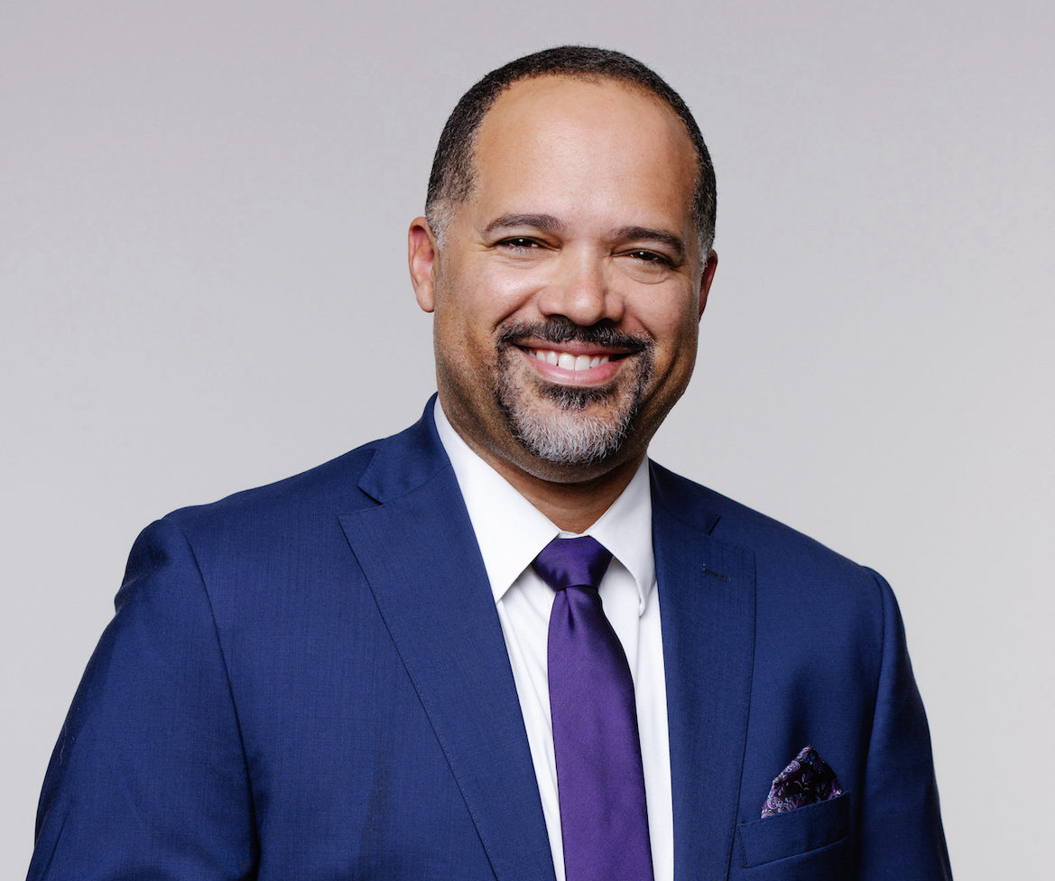 A smiling Black man wearing a blue business suit with a purple tie.