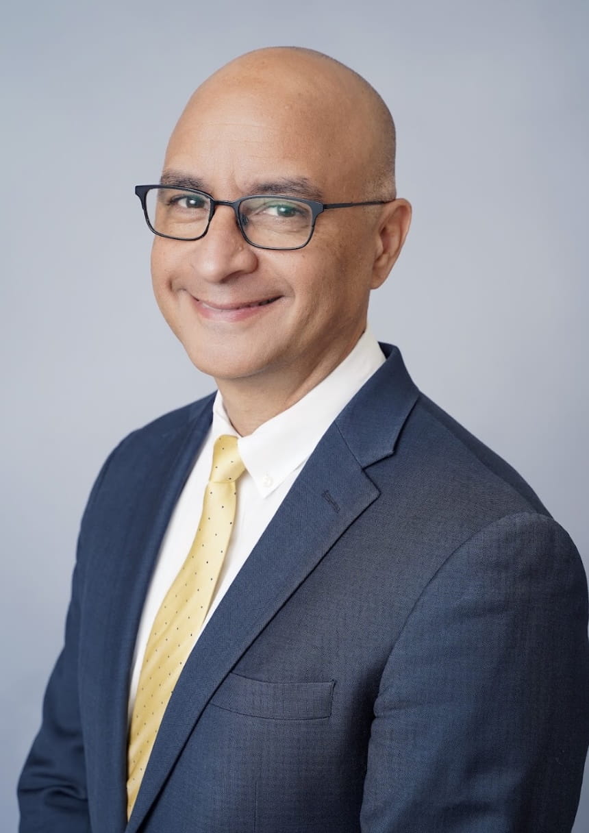 A man wearing glasses in a navy suit and sporting a golden-colored tie.