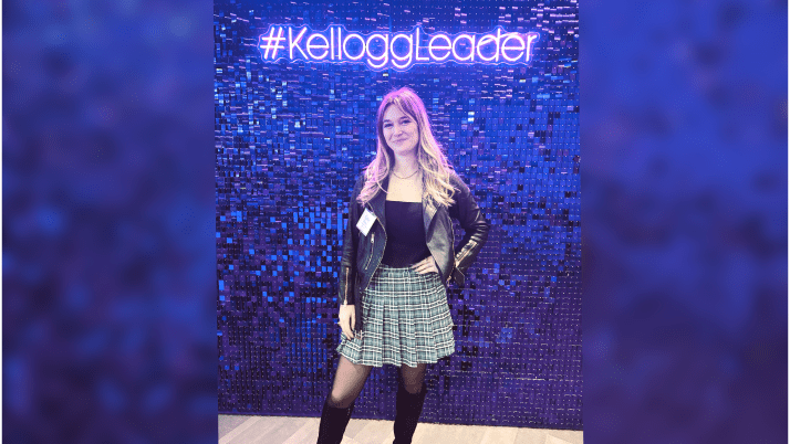 A female MBA student wearing a skirt and a leather jacket in front of a purple glittery wall