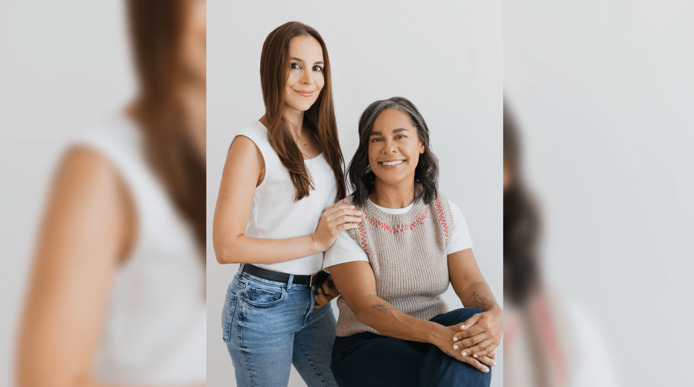 A brown-haired woman standing next to a woman sitting on a stool