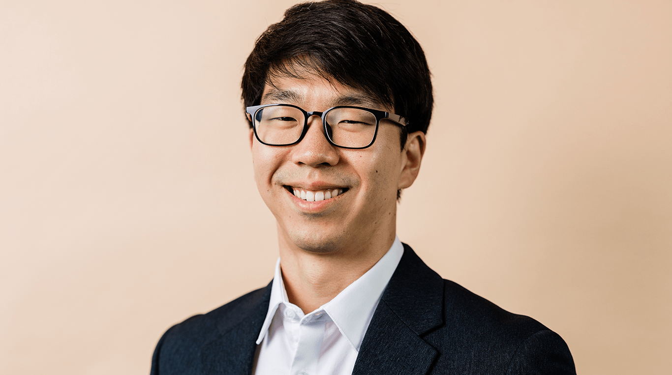 A smiling Asian man wearing glasses and a dark sport coat with a collared white shirt