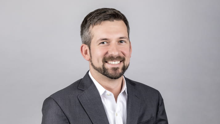 A white man smiling wearing a white collared shirt and blazer