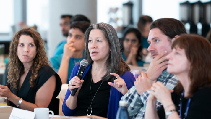 A woman wearing a black shirt with a purple cardigan and holding a mic in a group of people