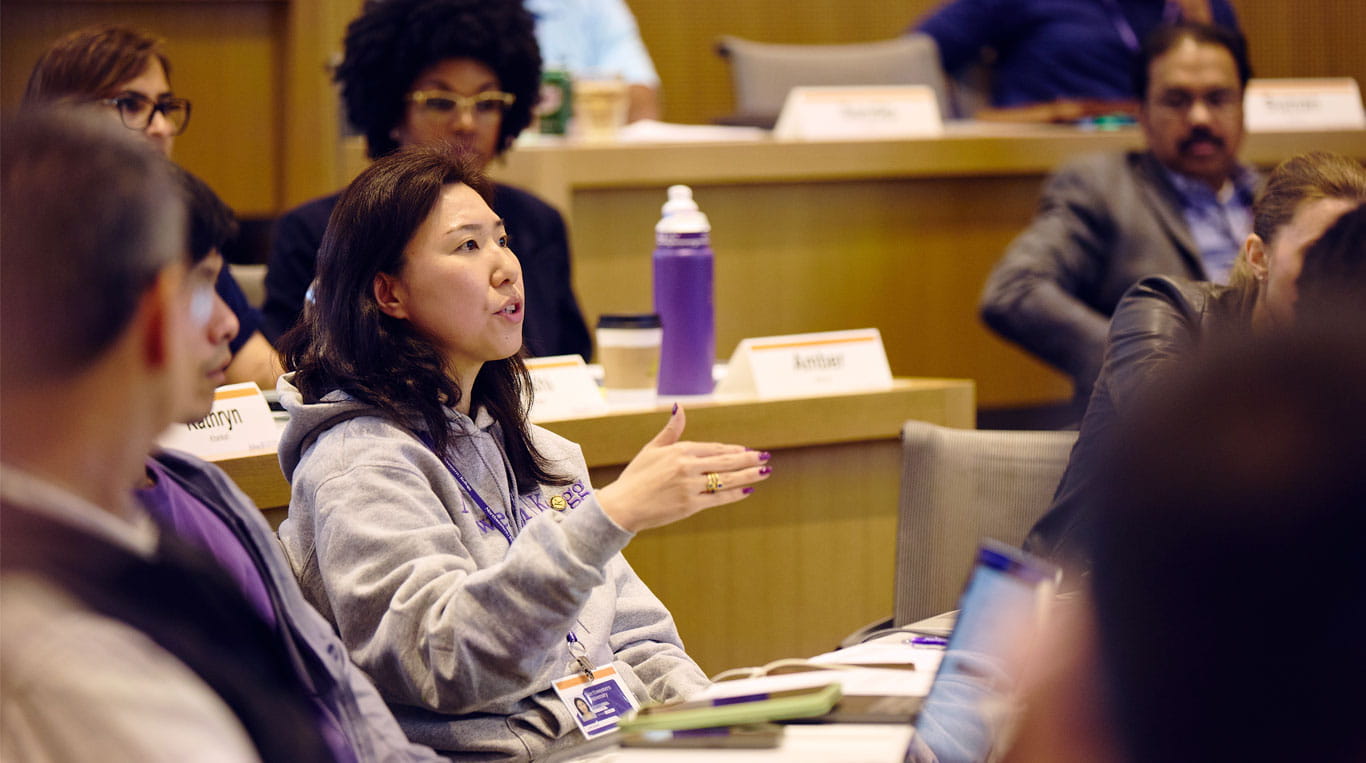 An Asian woman wearing a grey sweatshirt sitting in a classroom with other Kellogg students
