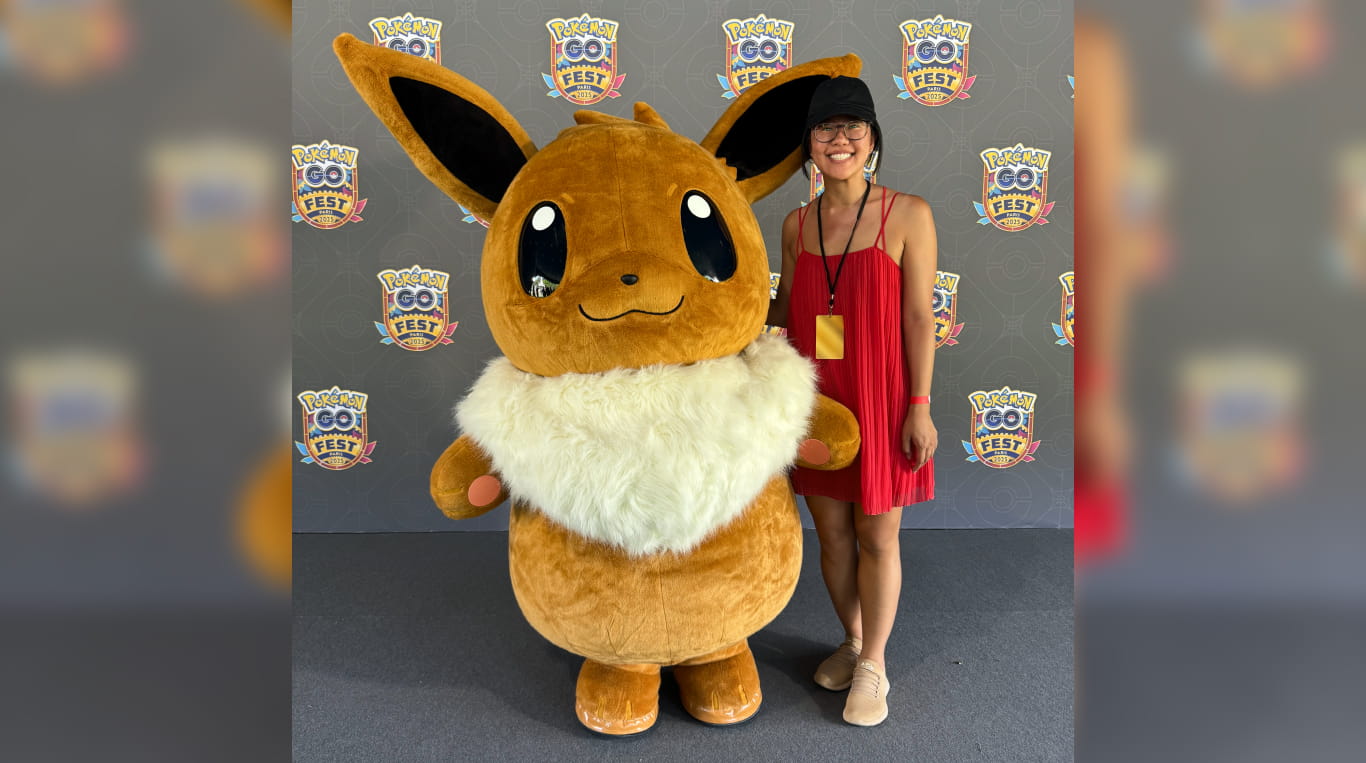 An Asian woman wearing a black hat and glasses stands next to an oversized Pokemon stuffed animal.