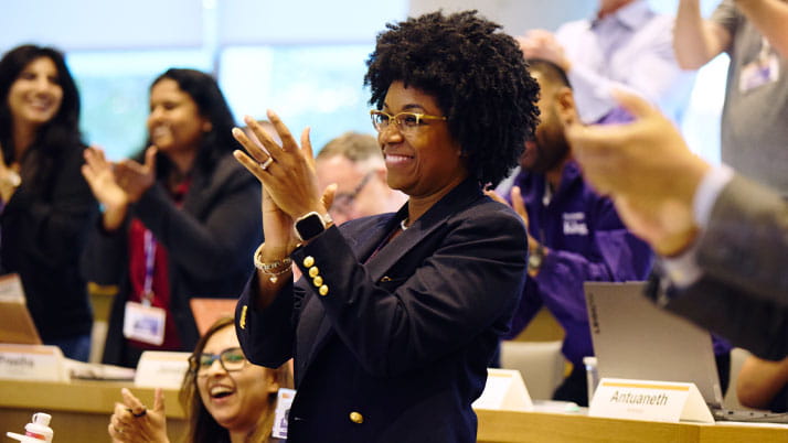 An African American woman wearing a blazer is standing and clapping.