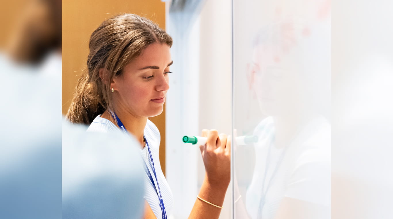 A woman with her hair in a low ponytail writing on a whiteboard.