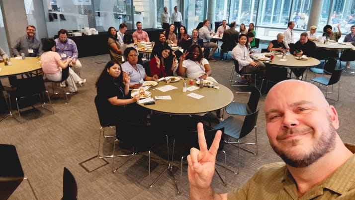 A white man making a peace sign with his hand and taking a group selfie.